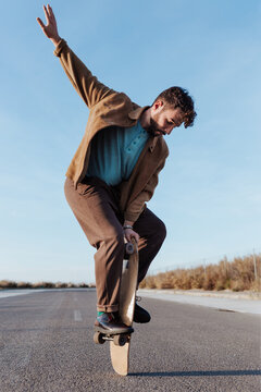 Full body young bearded male skater standing on edge of skateboard keeping balance while performing trick on asphalt road with hand raised and looking down
