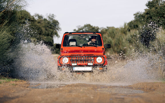 Unrecognizable Man Driving Red SUV Vehicle On Dirt Road With Water Puddle Among Green Trees In Summer Countryside