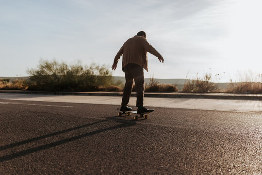 Full body back view of anonymous male skater in stylish wear riding skateboard along asphalt road in countryside