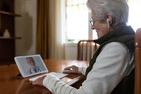 Side view of crop elderly female patient in TWS earbuds speaking with doctor on tablet during video call at home