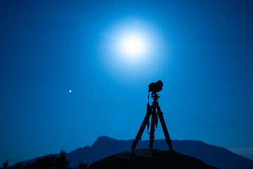 Professional photo camera with strap on tripod against mountain silhouette under colorful sky with sun in twilight