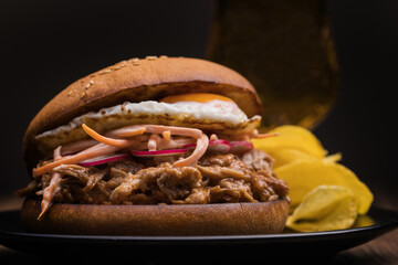 Closeup of appetizing hamburger made with pulled pork and fried egg with vegetables served with potato chips