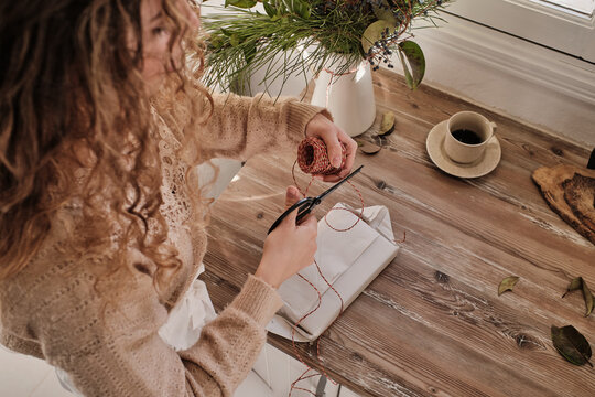 From above of creative female cutting rope and standing at table at home while wrapping and decorating gift