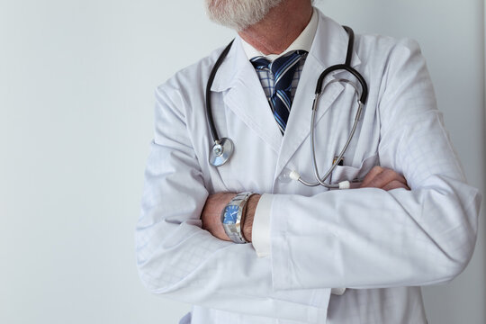 Crop senior male doctor with gray beard and folded arms in robe with stethoscope standing in clinic