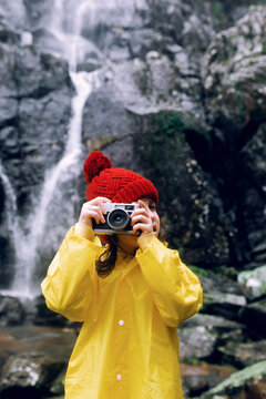 Anonymous teenager in raincoat taking photo on camera against rough mount with foamy cascade in daylight