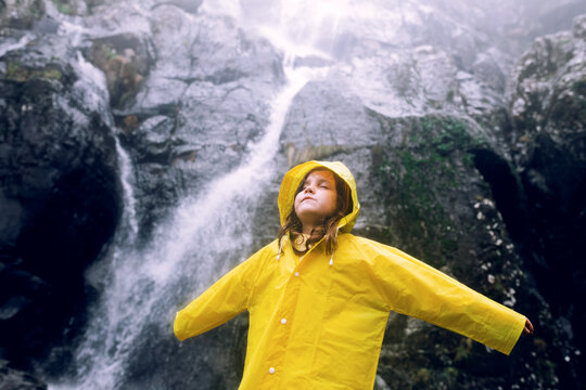 Female teenager in bright raincoat with eyes closed and raised arms standing against cascade with fast water flow on mount