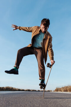 Full body young bearded male skater standing on edge of skateboard keeping balance while performing trick on asphalt road with hand raised and looking down