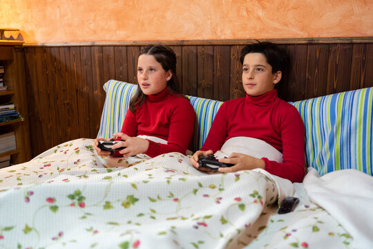 Cheerful teenage brother and sister sitting on bed and playing videogame while using gamepads and enjoying weekend at home