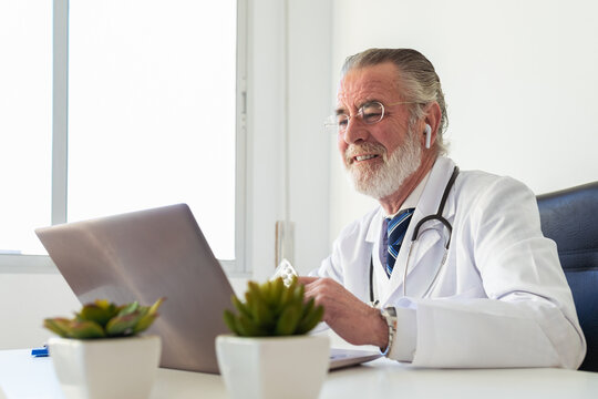 Elderly male doctor in earbud showing medications while talking on video chat against netbook during online health consultation in hospital