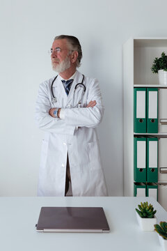 Senior male doctor with gray beard and folded arms in robe with stethoscope looking away in clinic