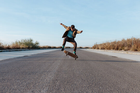 Full body young bearded skater in casual outfit jumping while performing kickflip on skateboard on asphalt road
