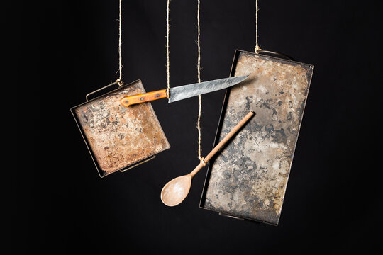 Weathered rustic metal baking sheets with knife and wooden spoon hanging on ropes against black background