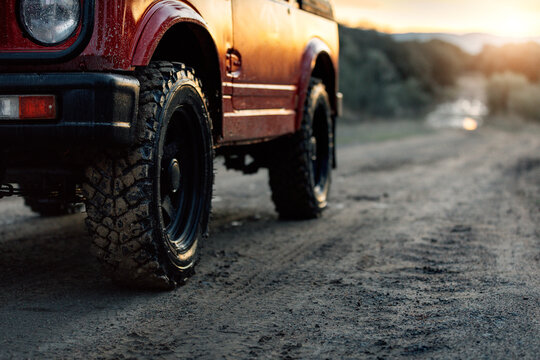 Cropped modern red off road car parked on dusty dirt road leading through forest in summer evening in countryside
