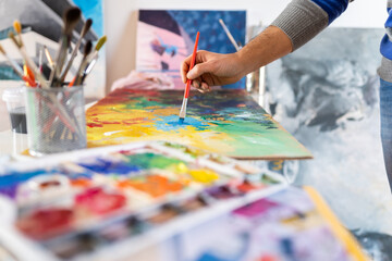 Crop unrecognizable male painter using professional brush during painting process on carton sheet near art tools in workroom