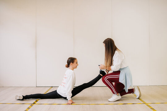 Side view of flexible girl sitting in split and stretching legs with help of dance trainer before lesson in spacious hall