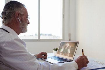 Side view of elderly male doctor in wireless earbud speaking with woman on video call on netbook in clinic