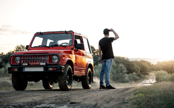 Full body back view of unrecognizable male adventurer standing near red off road car parked on dirt road among forest and looking away thoughtfully while enjoying summer journey in countryside