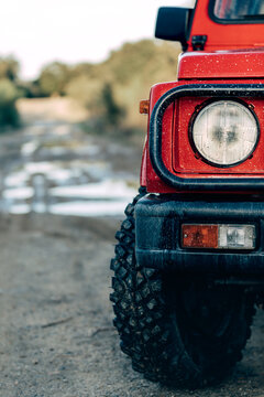 Closeup of headlight of red SUV vehicle parked on wet dirt path among green trees in countryside