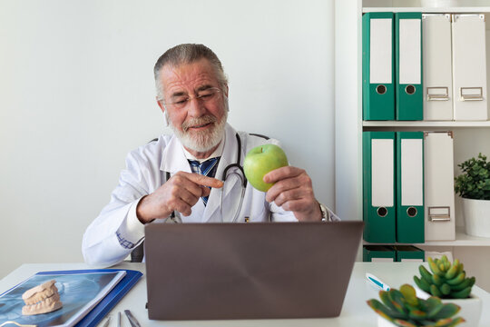Elderly male orthodontist showing green apple against portable computer while taking on video call in clinic