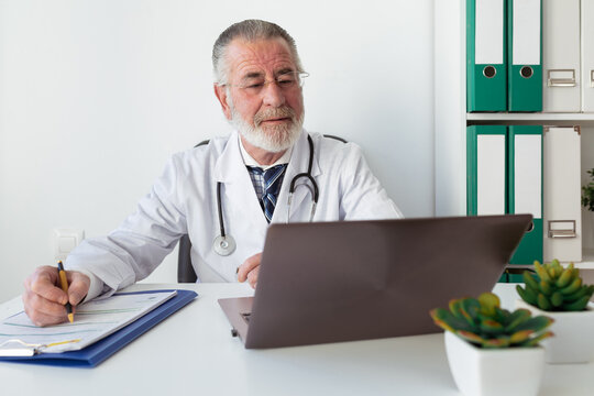 Senior male doctor in uniform writing memos on paper during online health consultation on portable computer in hospital