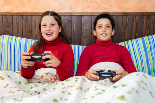 Cheerful teenage brother and sister sitting on bed and playing videogame while using gamepads and enjoying weekend at home