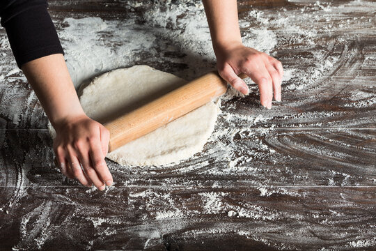 From above of crop anonymous female baker rolling dough on table sprinkled with flour while preparing bread in bakery kitchen