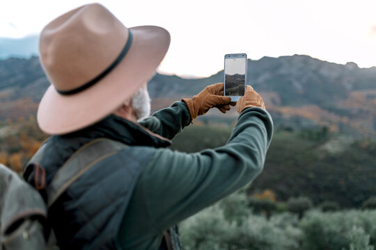 Back view of unrecognizable senior male explorer taking photo of mountainous landscape while using smartphone and enjoying vacation in Caceres