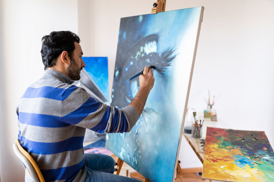 Side view of young bearded ethnic painter in casual wear siting in a chair painting with brush on canvas in art studio