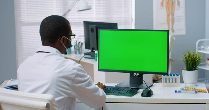 Back view of African American male physician sitting in hospital office and looking at green screen while working typing on computer. Medical concept, doctor browsing on monitor with chroma key
