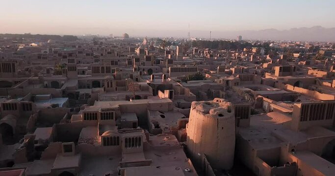 Aerial Fly Move Around the Old Desert City of Ardakan in Yazd in Morning Sunrise Time Over The Ancient Ruins of a Mudbrick Adobe Tower and winding Narrow Lanes in Hazy Day and Landscape of Mountains