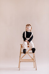 Fototapeta premium smiling little baby girl in beige jumpsuit and flower wreath on her head standing on a ladder in the studio. cute child isolated on beige background. Concept of the happy childhood.