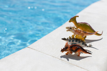 Four small toy dinosaurs stand in row near swimming pool on bright sunny day, copy space. Concept...
