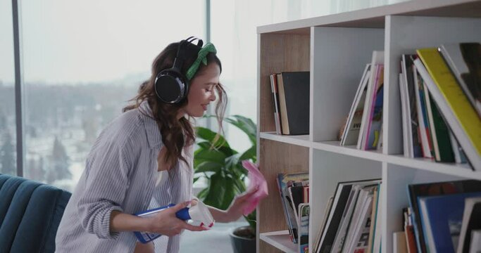 Caucasian beautiful excited woman staying home alone tyding the living room. Sexy attractive girl cleaning house into music wearing headphones.