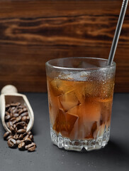 Low glass of coffee with milk, ice, straw and drops on the glass on black wooden background. Spoon with coffee beans. Selective focus on the front wall of the glass.