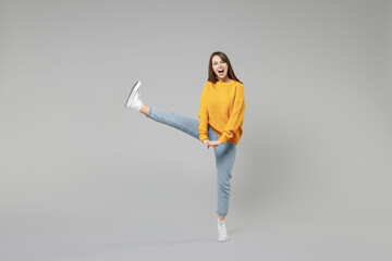 Full length of young smiling relaxed fun positive caucasian happy woman 20s wear knitted yellow sweater stand with fingers intertwined, raised up leg isolated on grey color background studio portrait