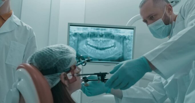 A female patient is sitting in the dentist chair in front of her is screen with scanned human jaw, she is wearing axiograph in front of her face ready for jaw tracking procedure.