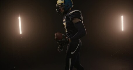 Strong rugby player with ball in studio. Muscular male rugby player in sportswear and helmet walking with ball in dark studio