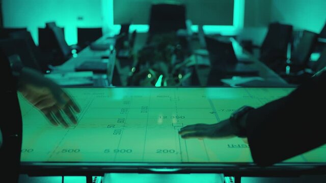Two men in black suits meeting at dark office room and using a modern, futuristic big touch screen tablet. Businessmen looking at map illuminated on digital table, discussing and talking . Slow Motion