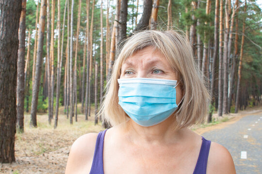 Beautiful Senior Woman In A Medical Mask Walks Outdoors In The Park On A Treadmill. Feeling Happy With Nature Background, Health Of The Elderly. Enjoying A Summer Day In The Nature, Freedom Concept.