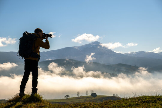 Silhouette of a backpacker photographer taking pictures of morning landscape in autumn mountains with digital camera.