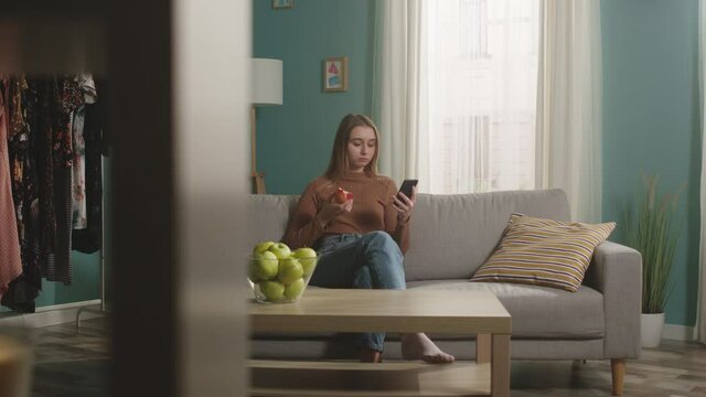 Young lady in blue jeans and brown sweater sits on beige sofa on window background, bit red apple and choked. Trying to cough. On table is glass jar with green apples. Camera dollies horizontally