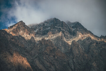 Mountain landscape with rocks in golden sunlight and low clouds. Awesome rocky wall with sharp top in gold sunshine in overcast weather. Atmospheric scenery with high rocky mountain pinnacle in clouds