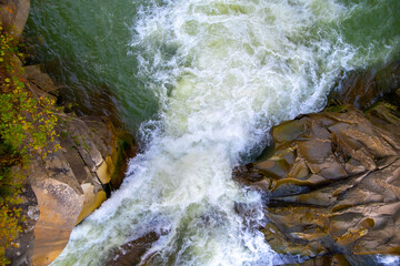 Aerial view of river waterfall with clear turquoise water falling down between wet boulders with thick white foam.