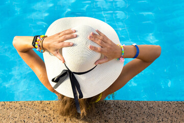 Fototapeta na wymiar Back view of young woman with long hair wearing yellow straw hat relaxing in warm summer swimming pool with blue water on a sunny day.