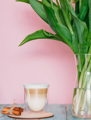 Cup of coffee with milk on a light pink background. Hot latte or cappuccino cooked with milk on a wooden table with copy space. Nearby is a transparent vase of flowers