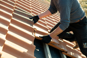 Closeup of worker hands installing yellow ceramic roofing tiles mounted on wooden boards covering residential building roof under construction. - obrazy, fototapety, plakaty