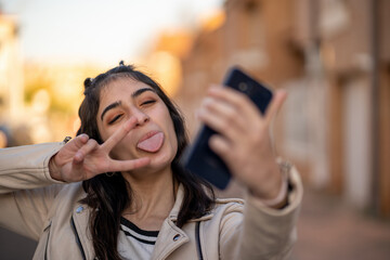 Mujer joven de pelo negro caucásica con camiseta de rayas negras, cazadora beige, se hace un selfie sonriendo con su smartphone sacando la lengua