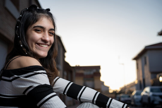 Mujer joven caucasica con camiseta de rayas negras y blancas, pelo negro, pantal&oacute;n vaquero gris y botas, sentada en un bordillo junto a la carretera. Moda. Estilo