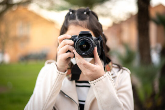 Joven mujer fotógrafa con una cámara réflex negra haciendo una foto al atardecer en un paisaje urbano, con cazadora blanca, camiseta de rayas, en un parque