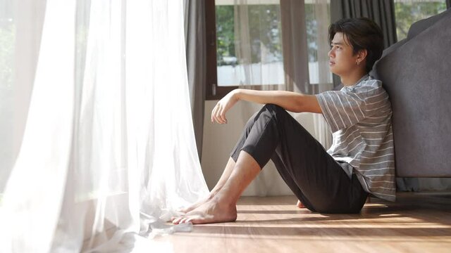 Depressed young Asian man sitting on the floor in living room with stressed and hopeless face. Loneliness sad guy being alone feeling bad and negative thinking. Mental health and life problem concept